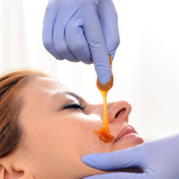 Gloved hands applying orange facial wax to a woman’s nose during a beauty treatment