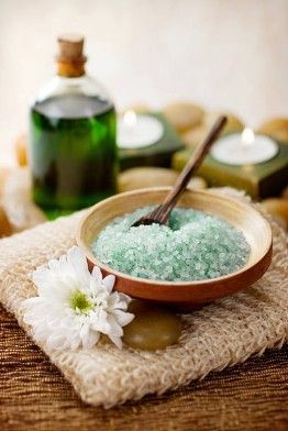 Spa salt in a bowl with a wooden spoon, white flowers, and a green bottle on a woven mat
