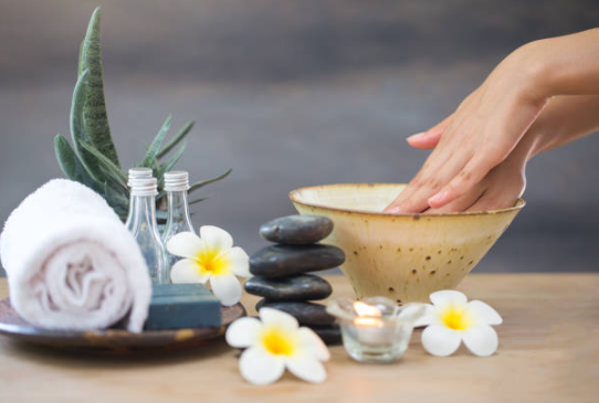 Spa setup with hand in bowl, stacked stones, candles, rolled towel, and white flowers on a wooden table