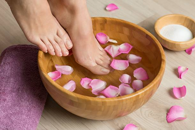 Feet soaking in a wooden bowl with pink flower petals, next to a towel and salt dish