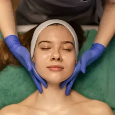 Gloved hands massage a woman’s face as she lies with eyes closed on a treatment bed.
