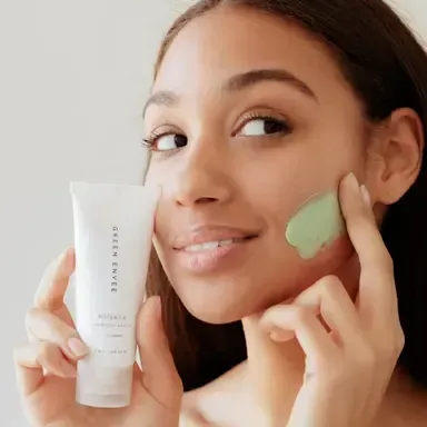 Woman applying green cream to her cheek beside a white skincare tube