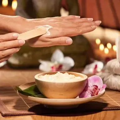 Hands receiving a spoonful of cream over a bowl on a spa tray with flowers and candles