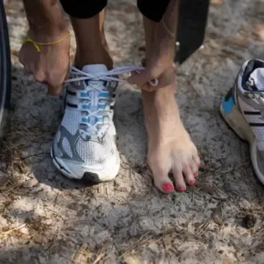 A person tying the laces of a blue-and-white running shoe beside a bare foot on rocky ground.