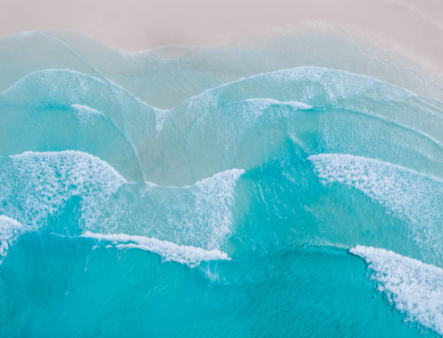 Turquoise ocean waves rolling onto a pale sandy shore, viewed from above.
