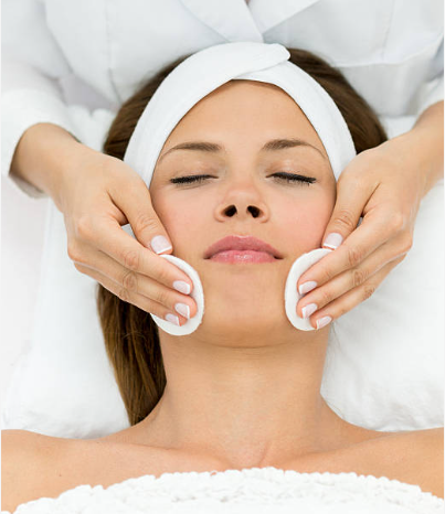 Spa therapist gently massages a woman's face with cotton pads, eyes closed and towel wrapped around her head