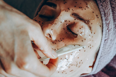 Facial mask being applied with a brush to a relaxed person lying down with eyes closed, wrapped in a towel
