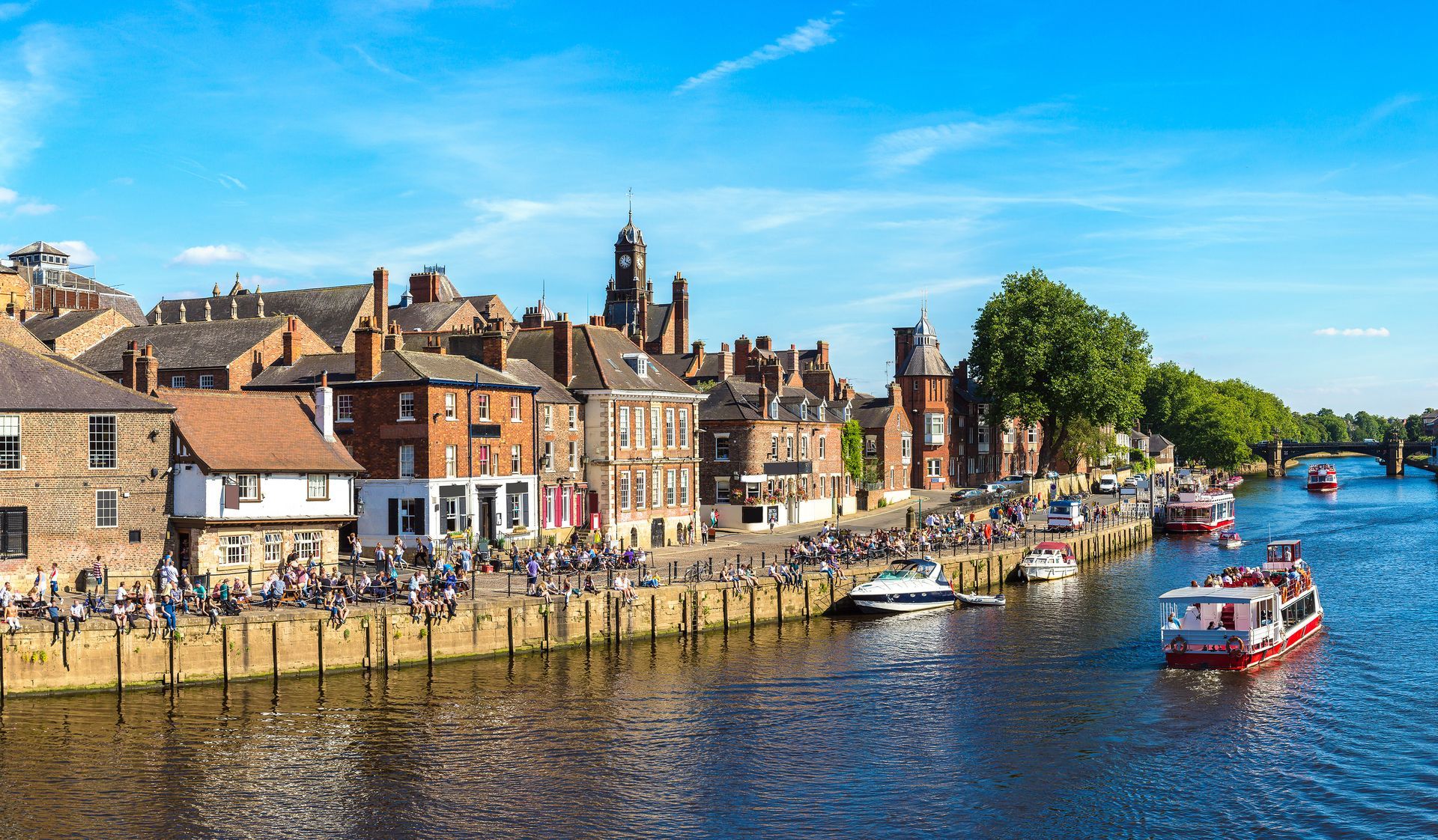 River scene of York, England, with buildings and people along the riverbank and boats on the water under a blue sky.