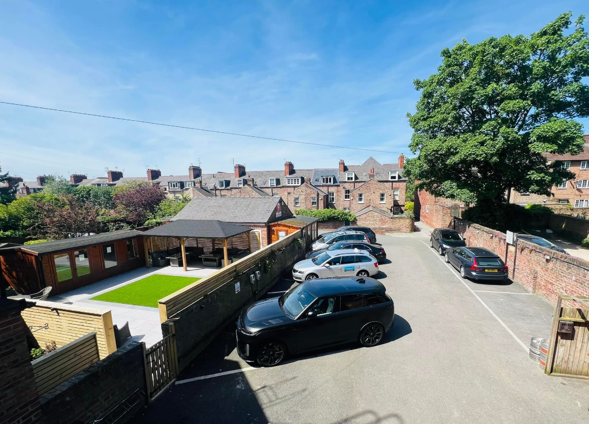 Aerial view of a row of brick buildings with cars parked in front. Green trees and a small garden are also visible.
