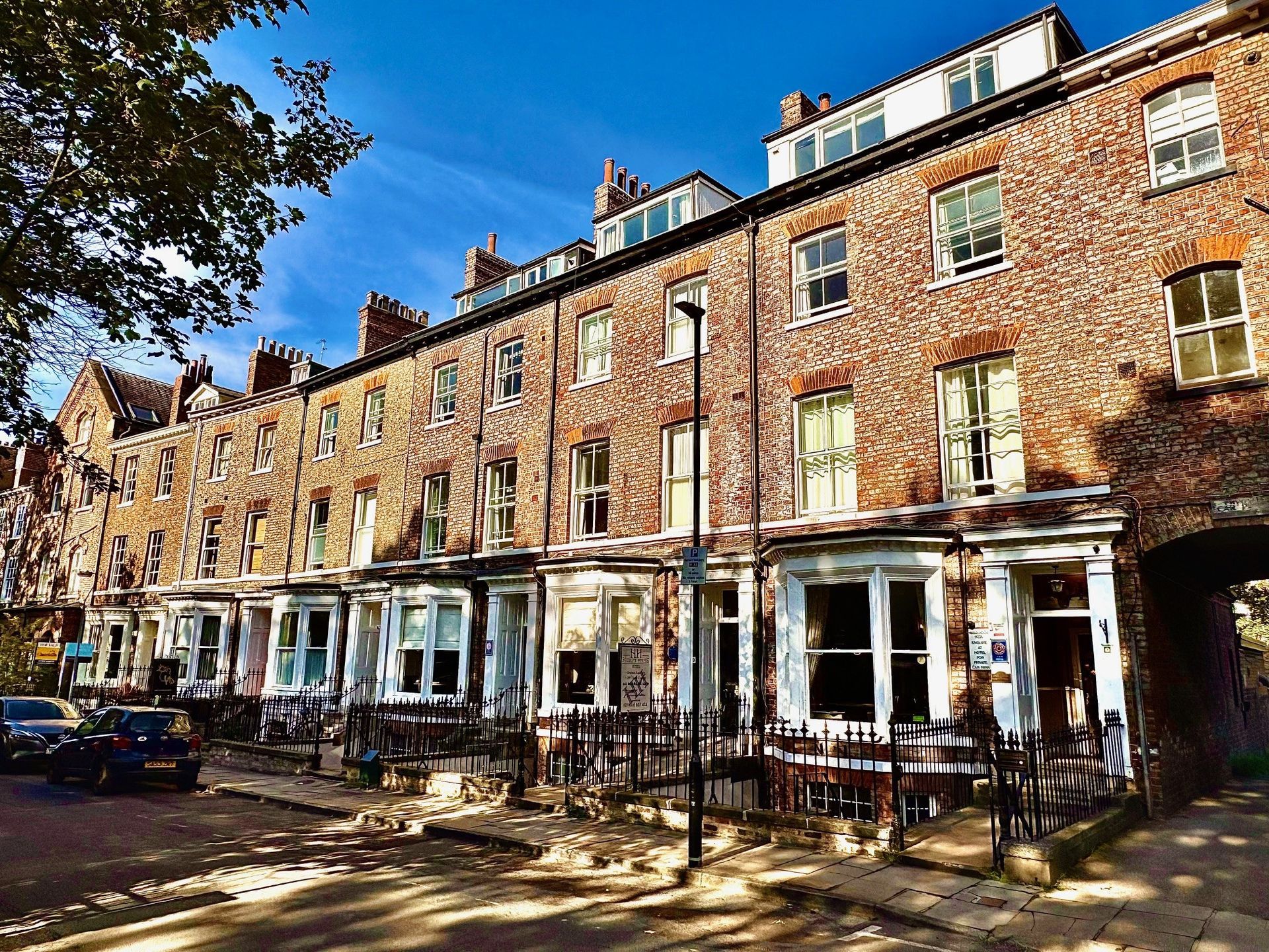 Row of brick buildings, one with a white-framed bay window and tables outside. Cars line the street.