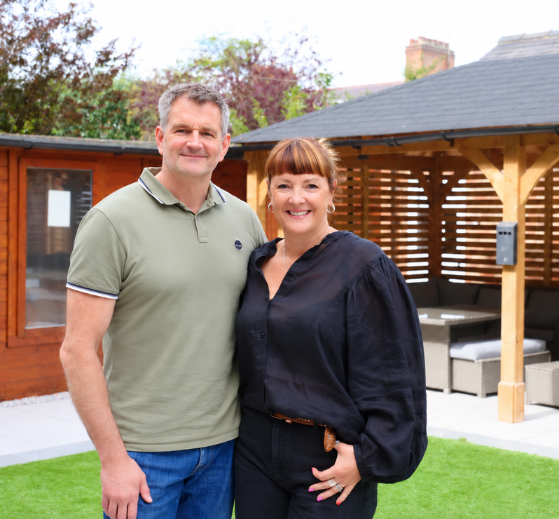 Man and woman smiling, posing in a backyard. Man wears olive green polo and jeans. Woman wears black top and pants.