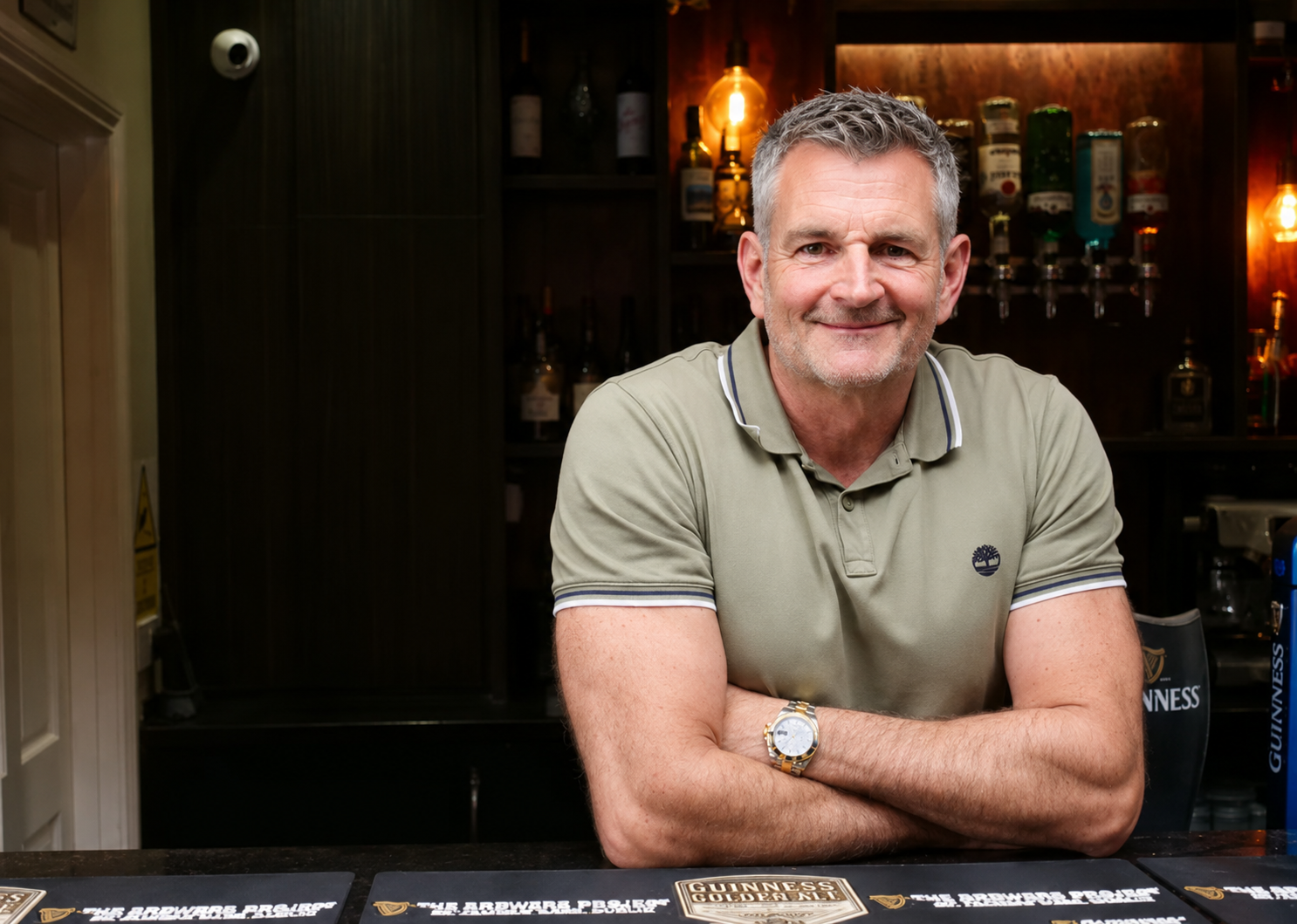 Man with gray hair, arms crossed, smiling at a bar. He wears a green polo shirt.