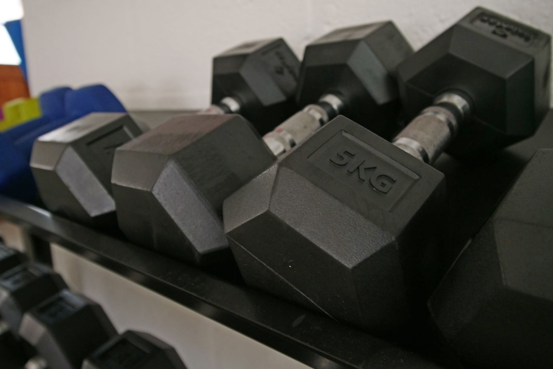 Black hexagonal dumbbells on a black rack in a gym. The weight 5KG is visible on one.