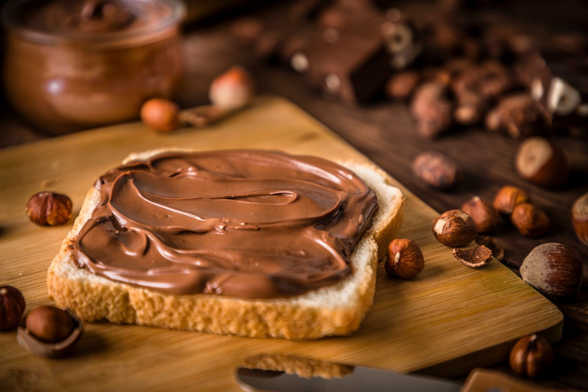 Una fetta di pane generosamente spalmata con crema al cioccolato e nocciole su un tagliere di legno, circondata da nocciole sparse.