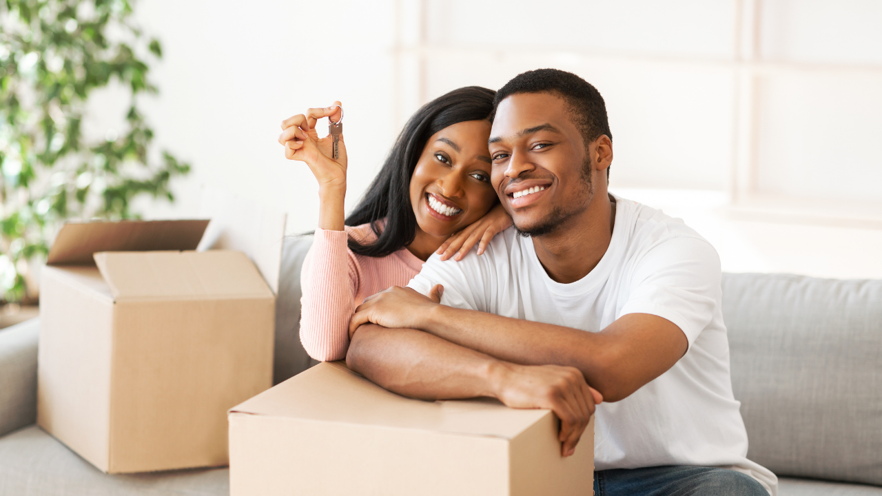 A smiling couple sits on a couch behind cardboard boxes, holding a key in a new home.