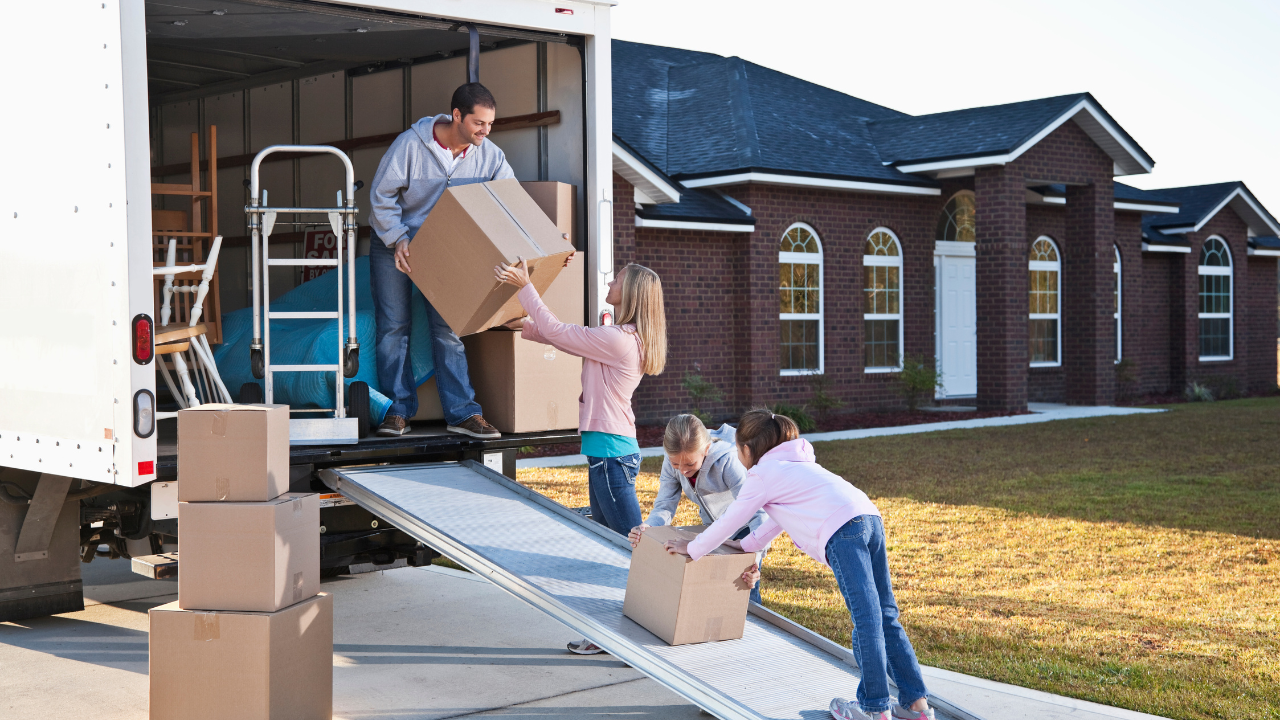 Family moving boxes from a truck into a brick house; father and children assisting.