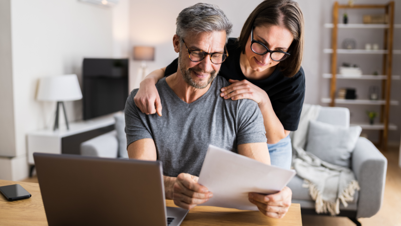 Couple reviews paperwork at home. Man sits at desk with laptop; woman looks over his shoulder.