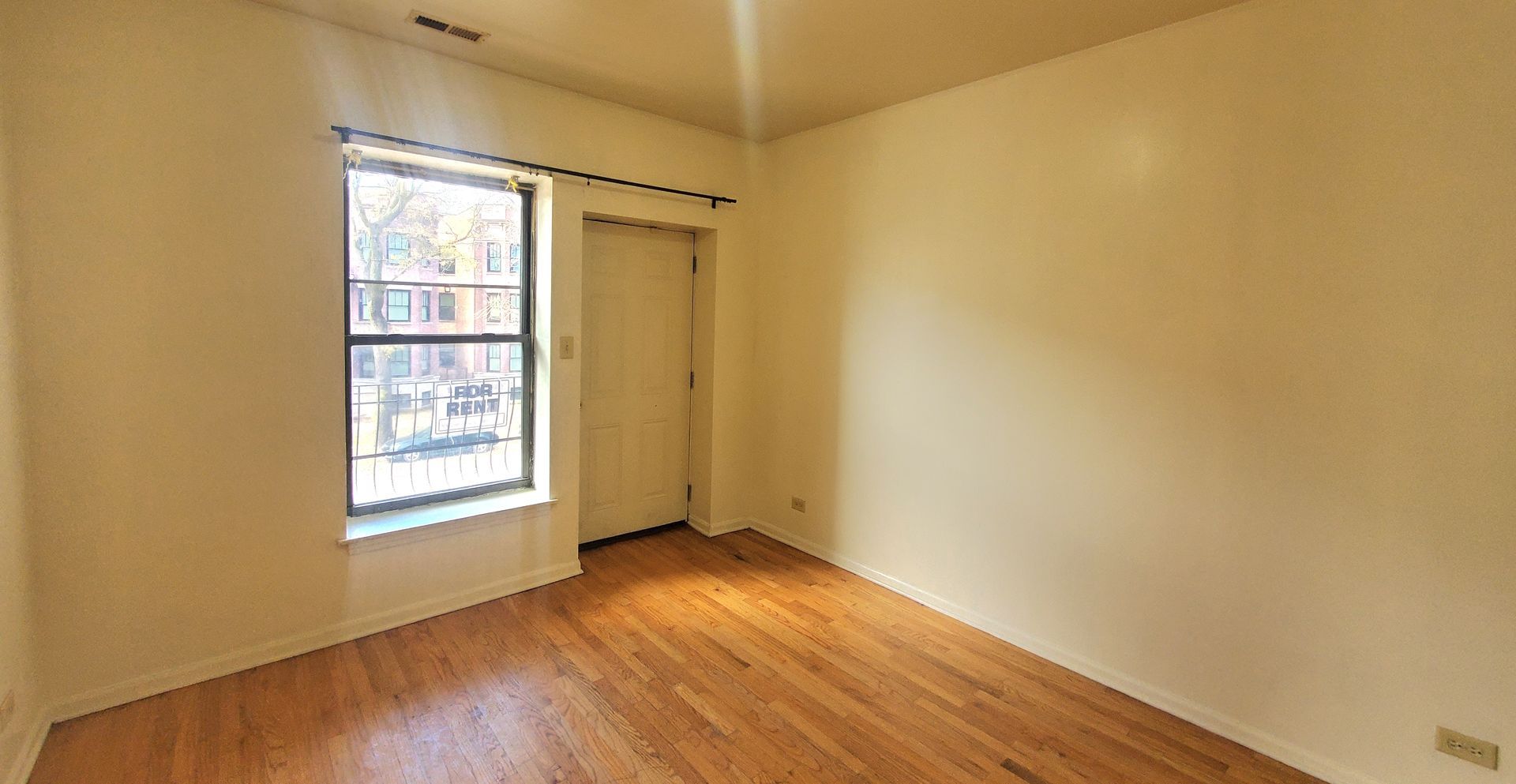 An empty room with light-colored walls, light wood flooring, a window, and a closed door.