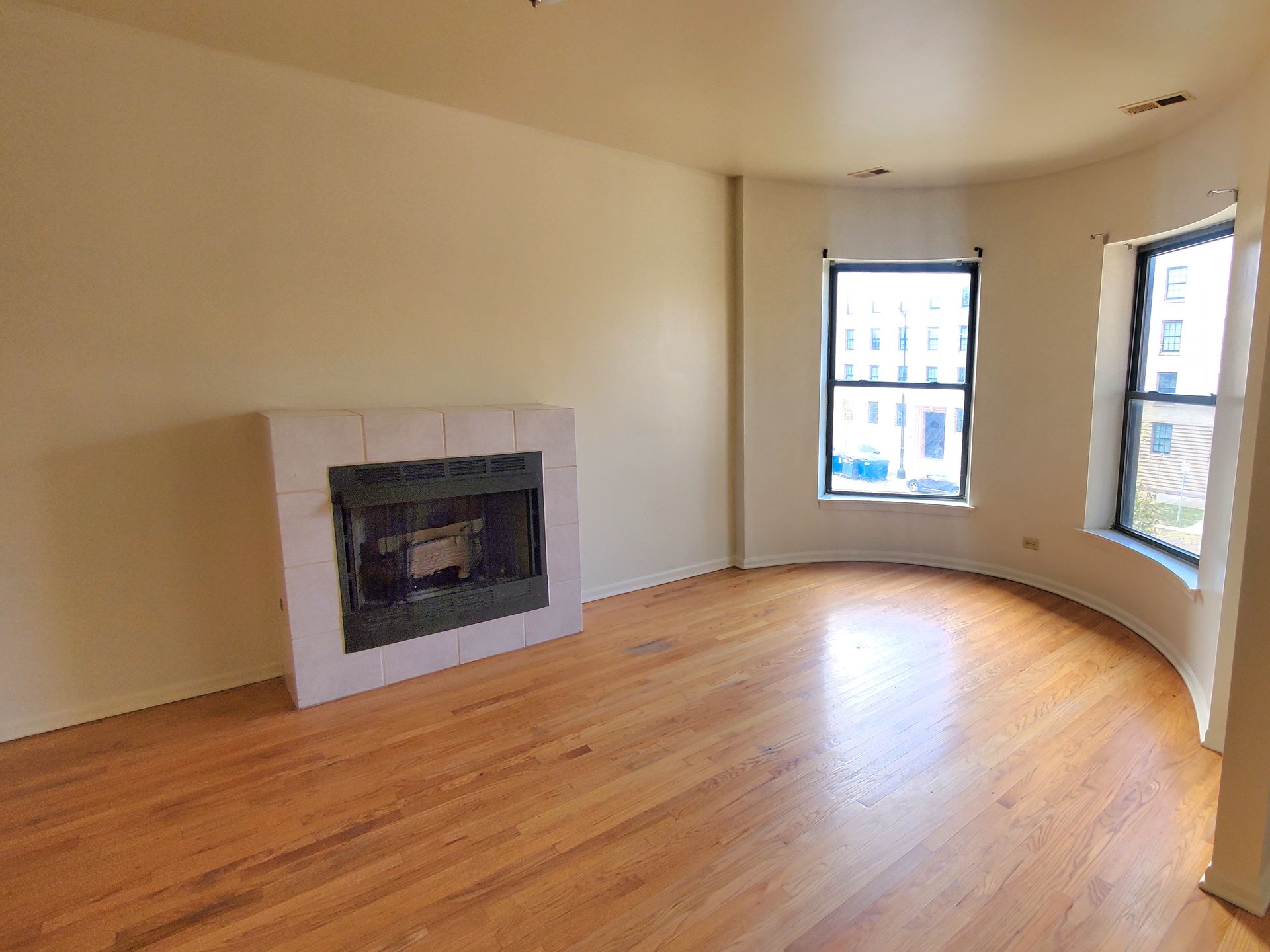 A bright, empty living room with light hardwood floors, a tiled fireplace, and two windows in a curved bay wall.