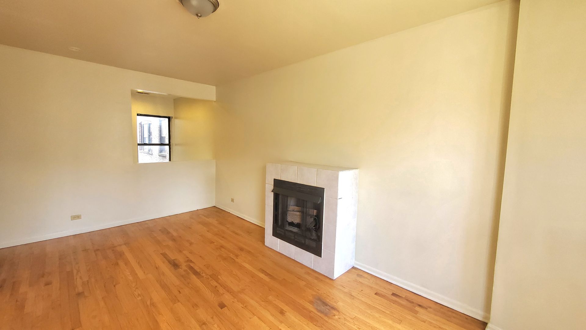 Empty room with light wood floors, beige walls, a white-framed fireplace, and a small window in a recessed alcove.