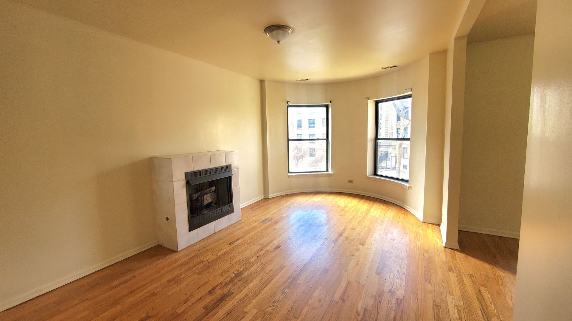 Empty living room with light hardwood floors, beige walls, and a fireplace in front of a curved wall with two windows.