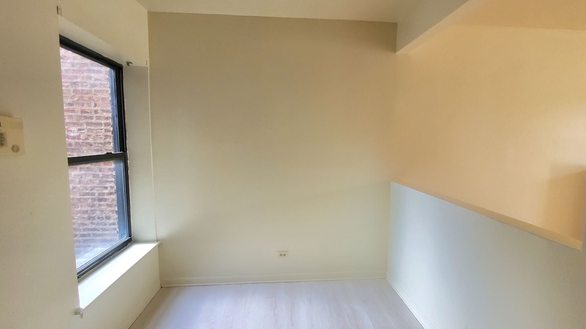 A small, empty room with light beige walls, a window overlooking a brick building, and white flooring.