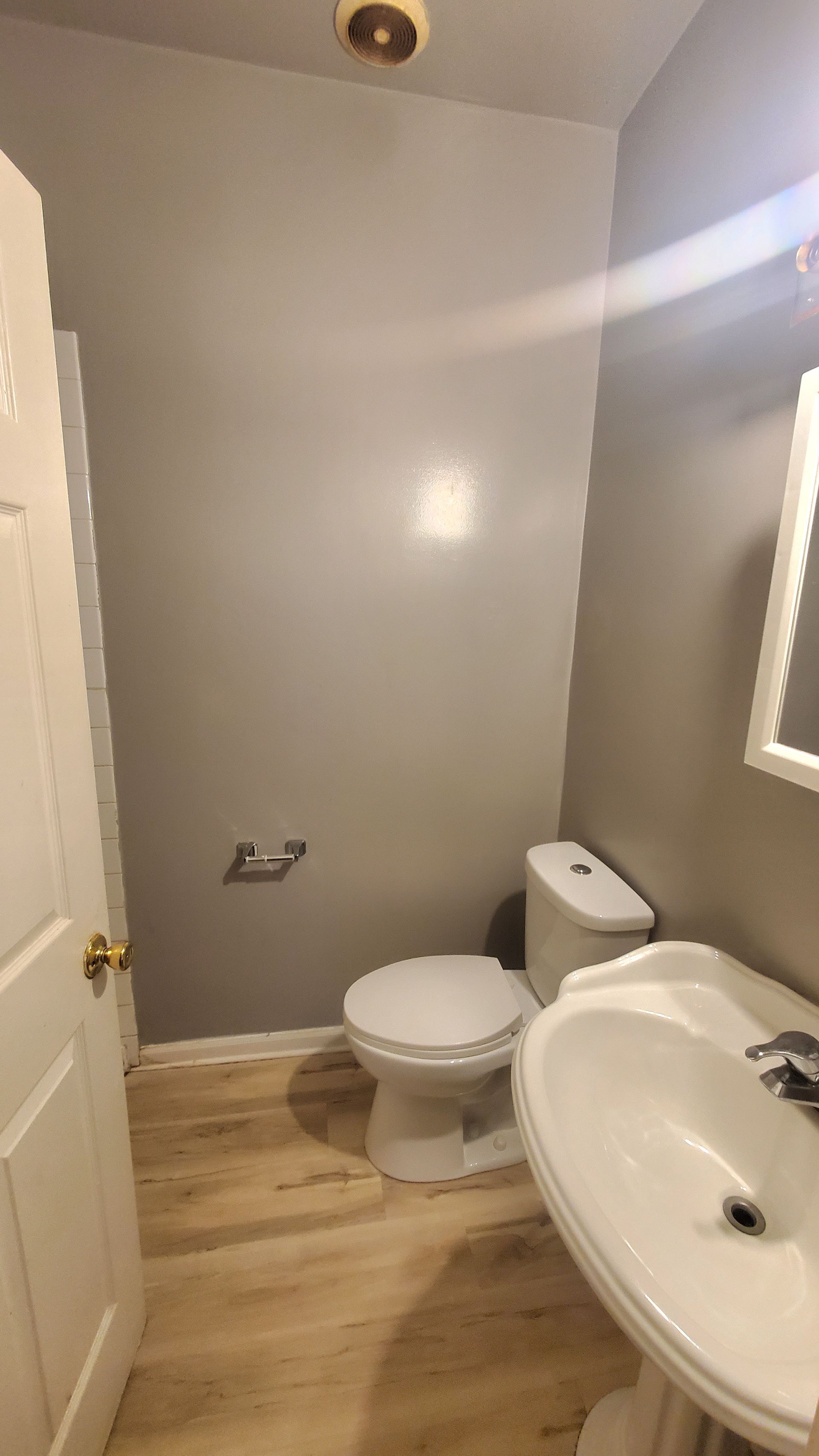 A small bathroom with light gray walls, light wood flooring, a white pedestal sink, and a white toilet.