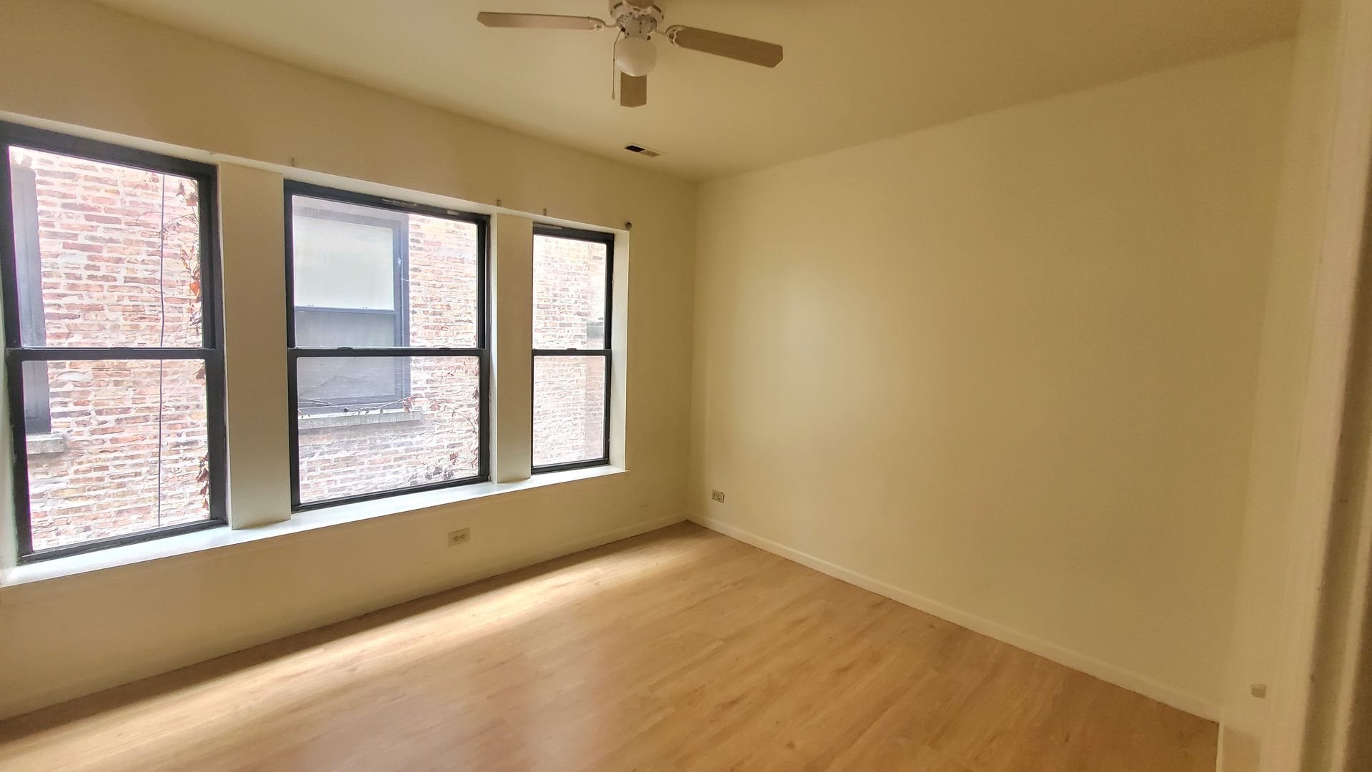Empty bedroom with beige walls, light wood flooring, a ceiling fan, and three large windows overlooking a brick building.