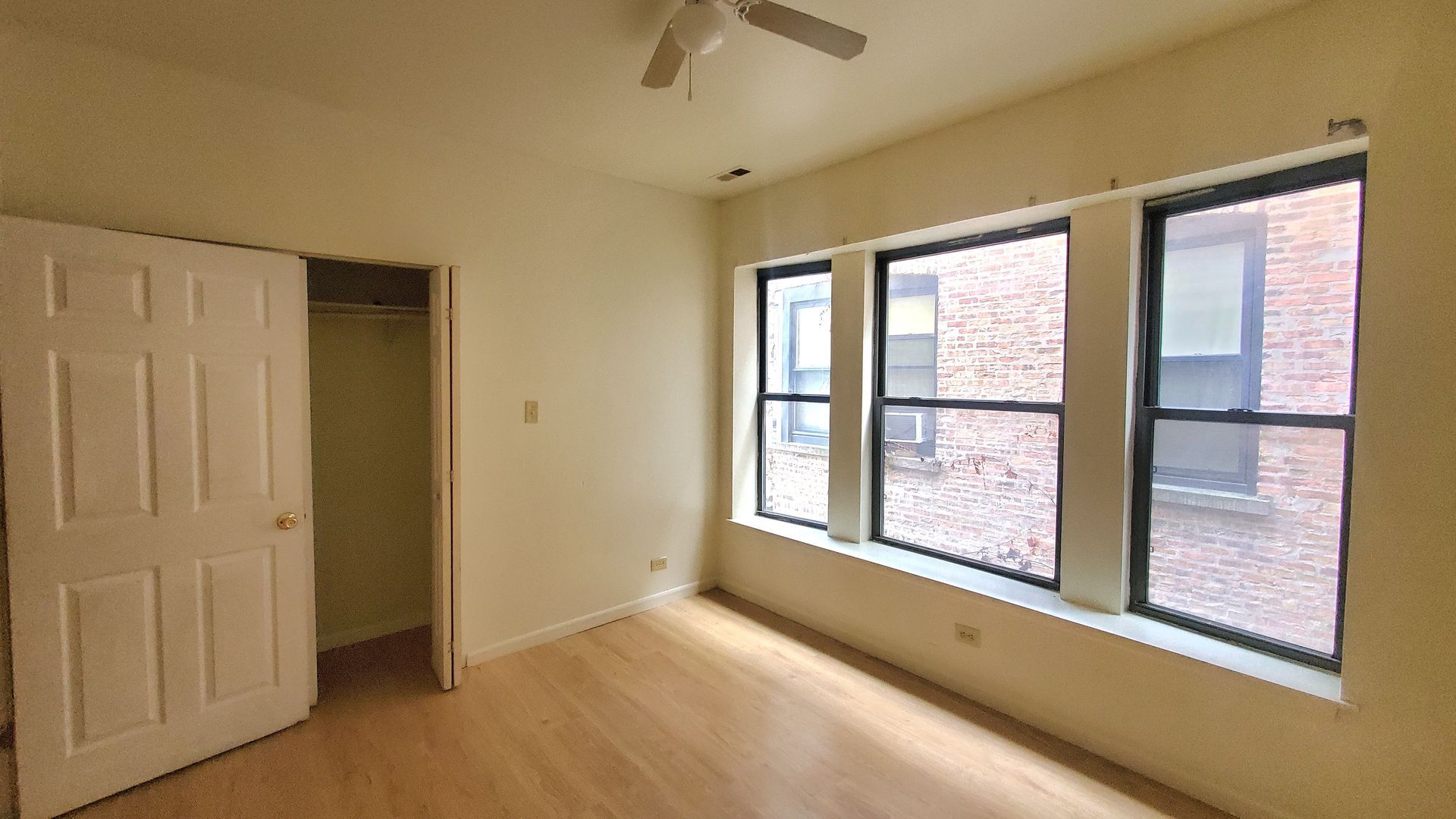 An empty bedroom with light-colored flooring, beige walls, an open closet door, and three large windows overlooking brick.