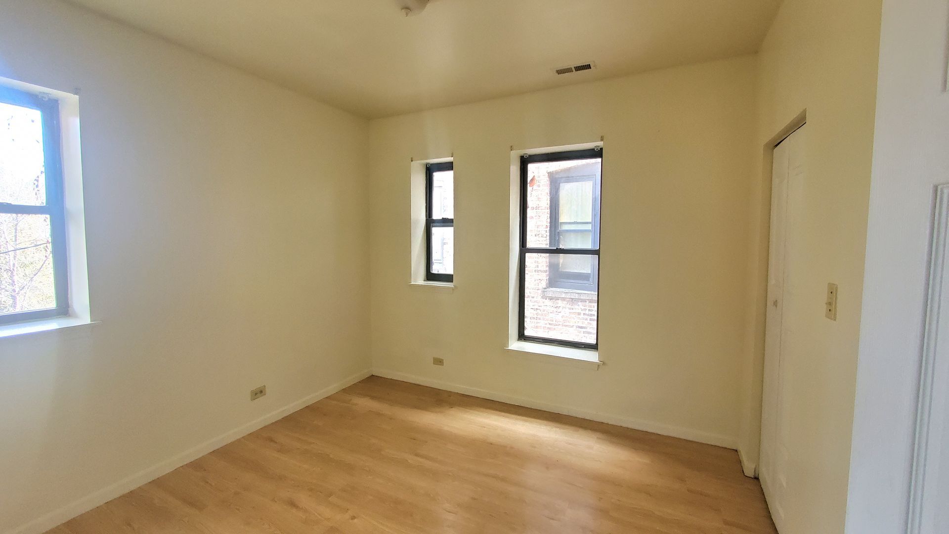 An empty bedroom with cream-colored walls, light wood flooring, three windows, and a closed white door on the right.