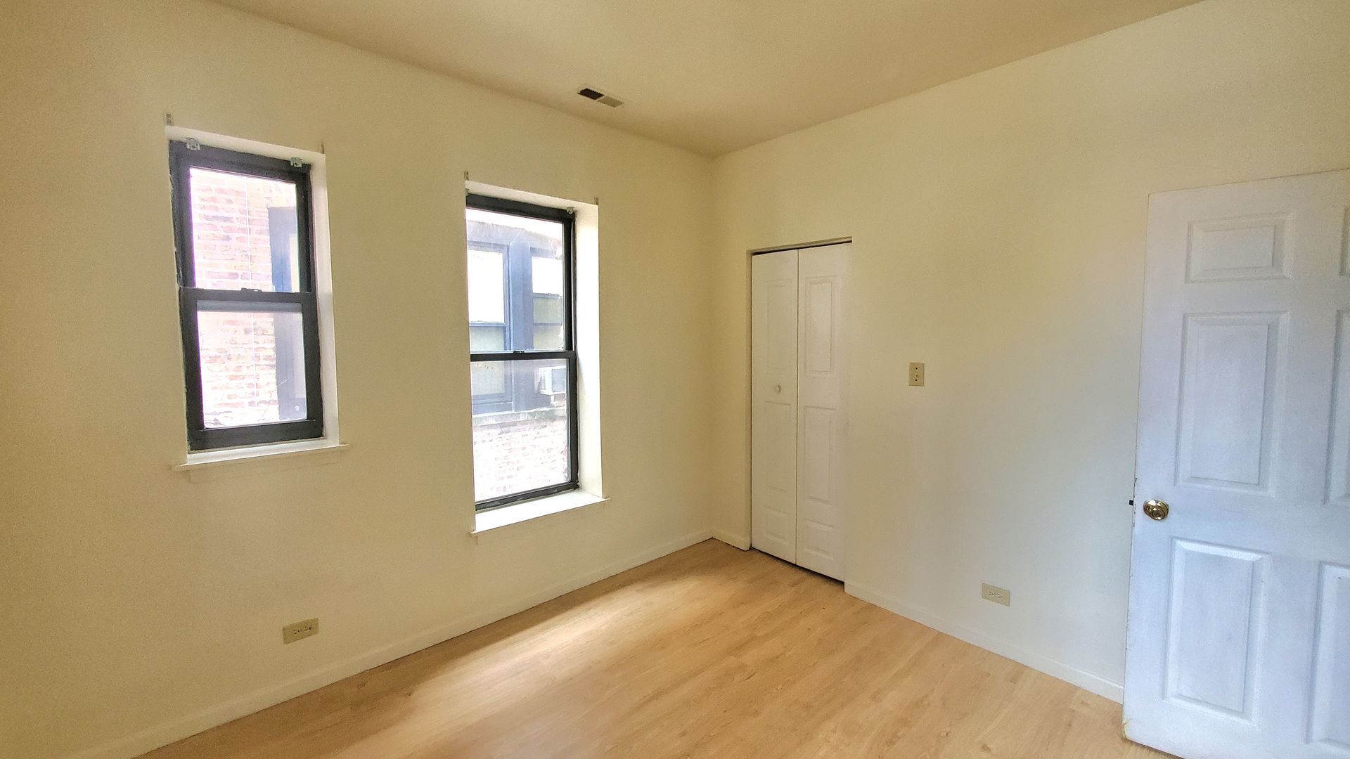 An empty room with light-colored walls, light wood flooring, two windows, a white door, and a white closet door.