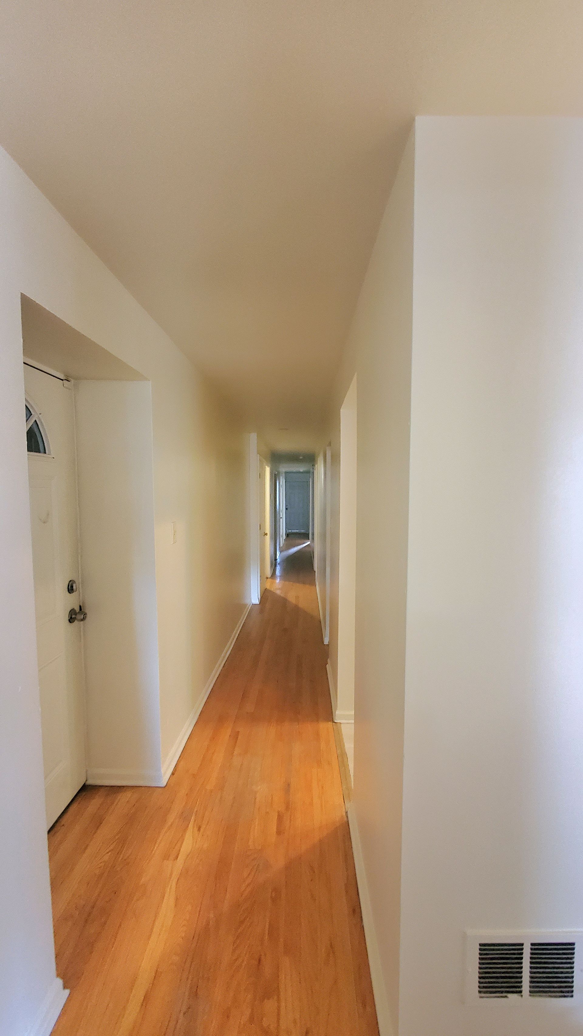 A long, narrow indoor hallway with light wood flooring and plain white walls, leading to a doorway at the far end.