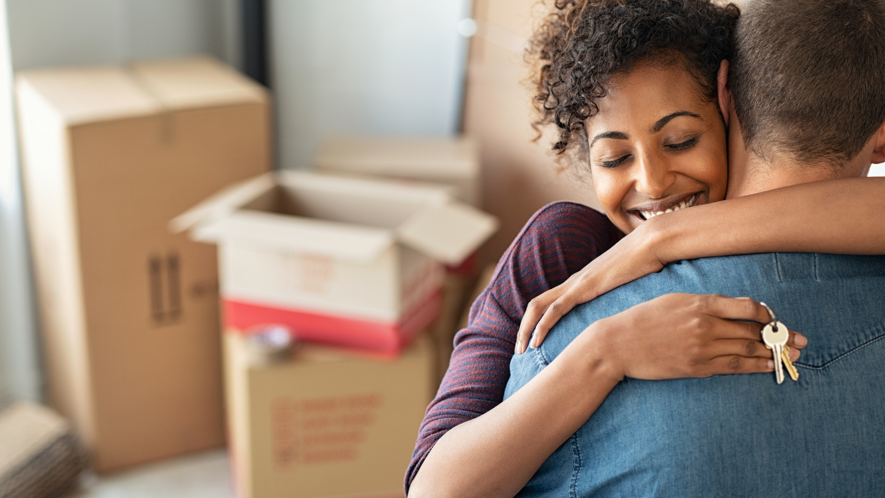 A couple embraces in an apartment filled with cardboard moving boxes, holding a set of house keys.