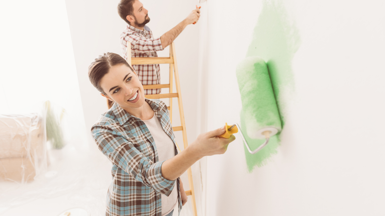 A person paints a wall with a green roller while another person on a ladder paints an upper section in a bright room.