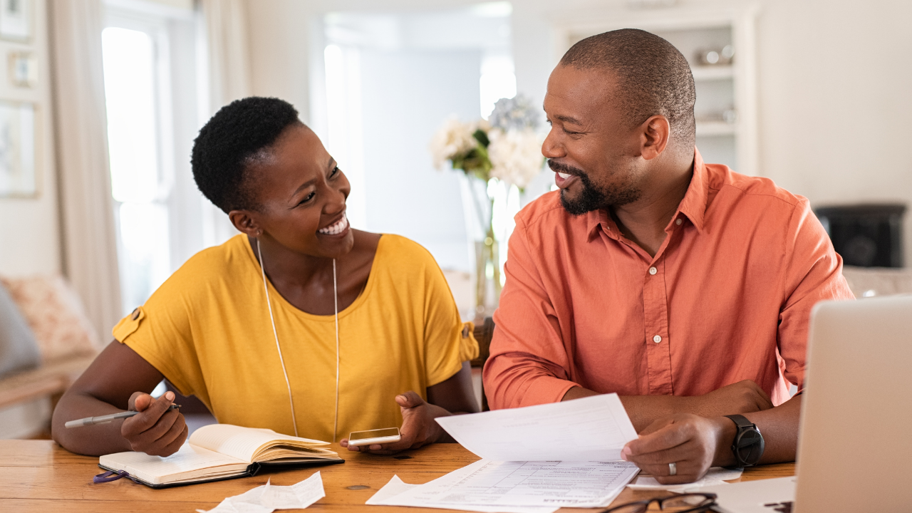 Couple smiling while reviewing paperwork at a table, notebook and laptop present.