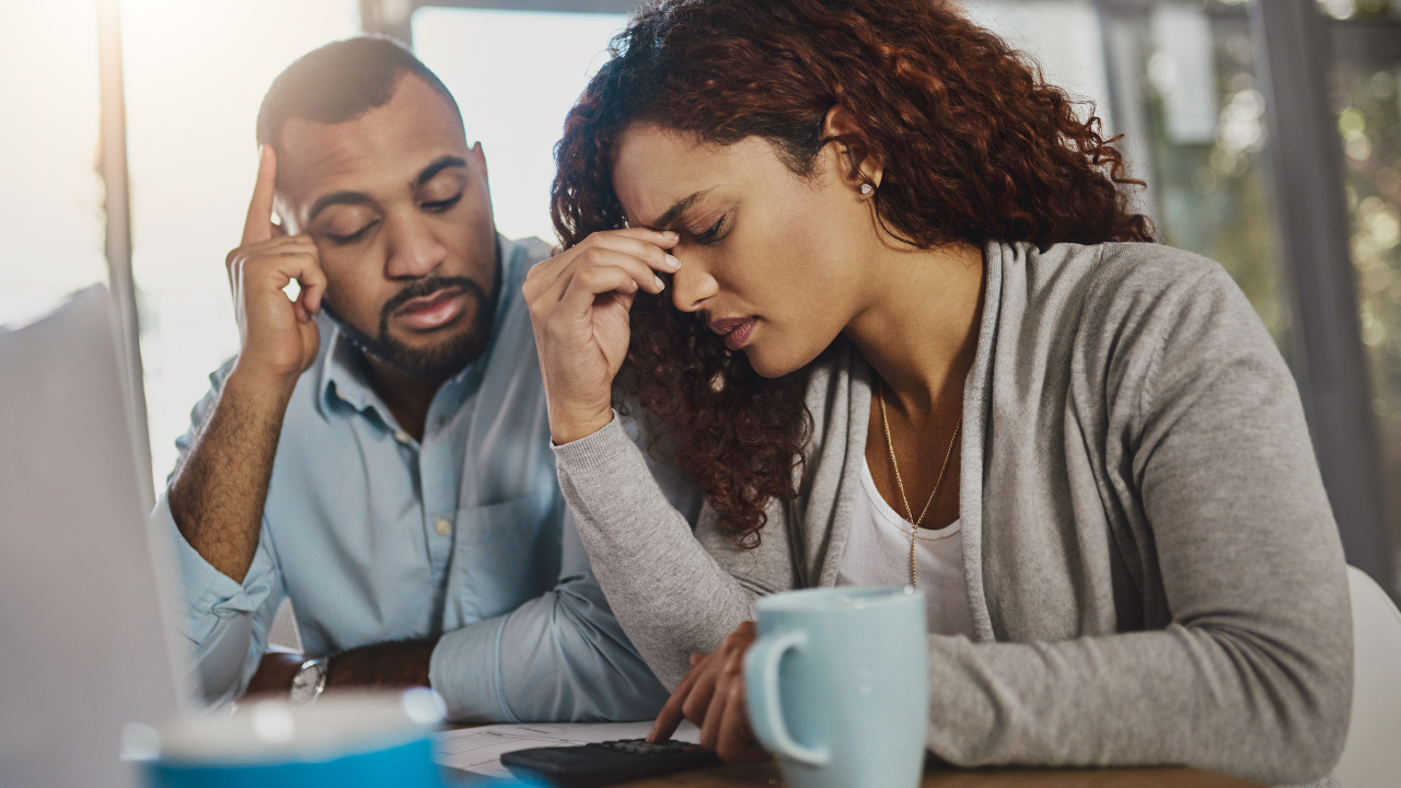 Couple looking stressed, calculating bills. Man has hand on his head, woman holding her forehead.