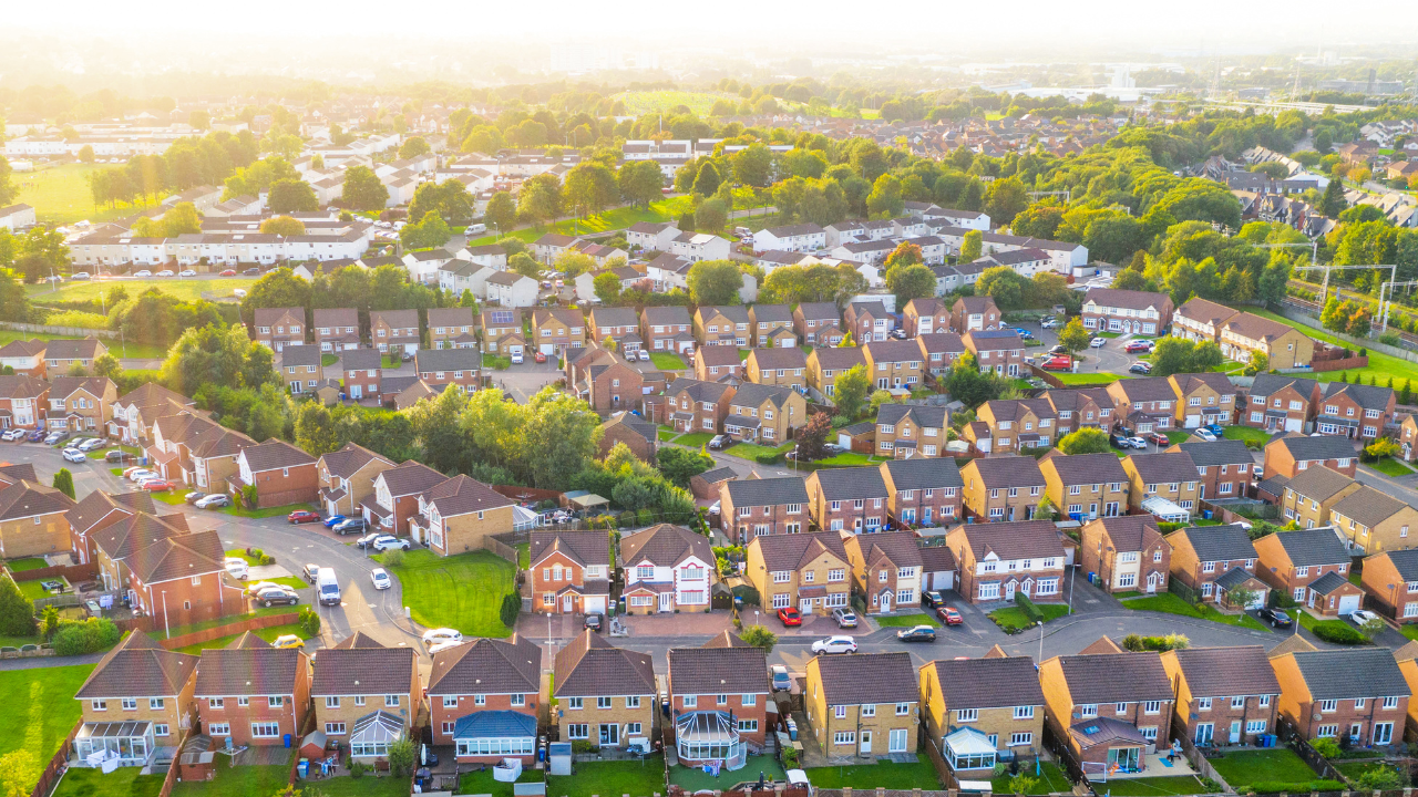 Aerial view of a suburban neighborhood with rows of houses, trees, and green spaces under a warm, sunny sky.