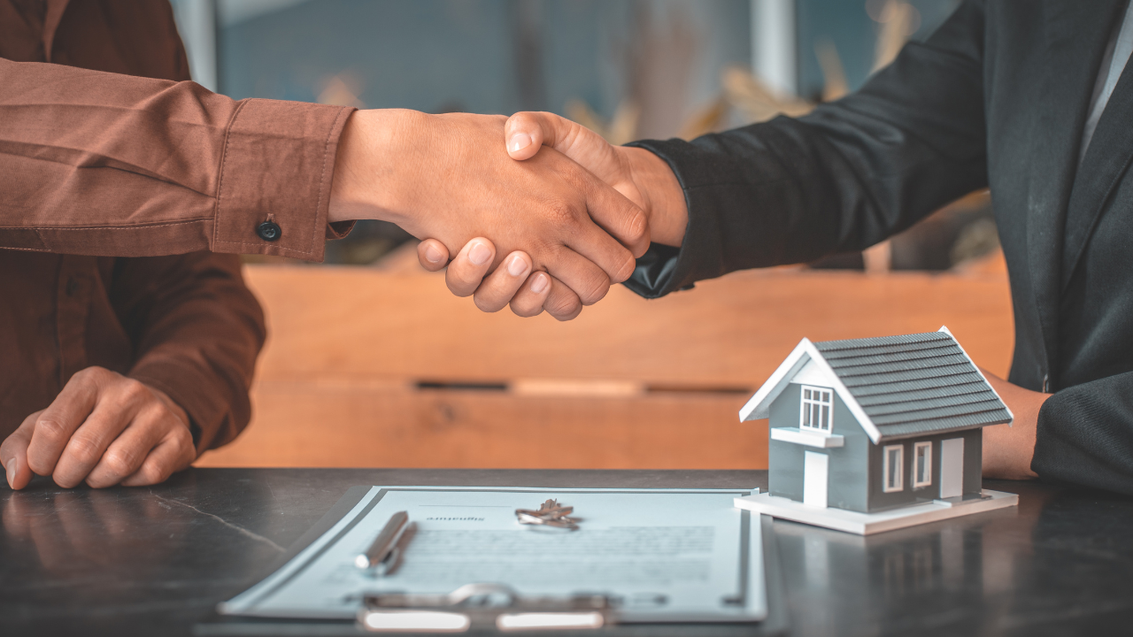Two people shaking hands over a house model, paperwork, and keys, likely closing a real estate deal.