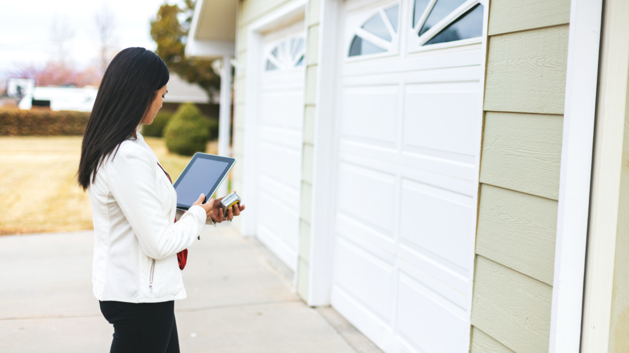 Woman in white jacket, black pants holding tablet by a garage door, outside a house.