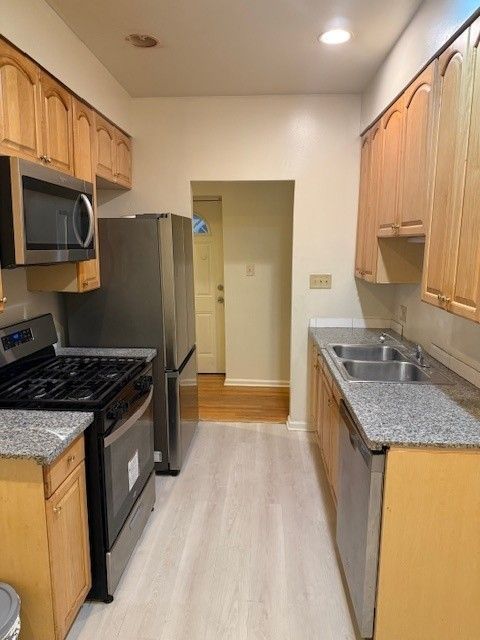A galley kitchen with light wood cabinets, stainless steel appliances, speckled countertops, and wood flooring.