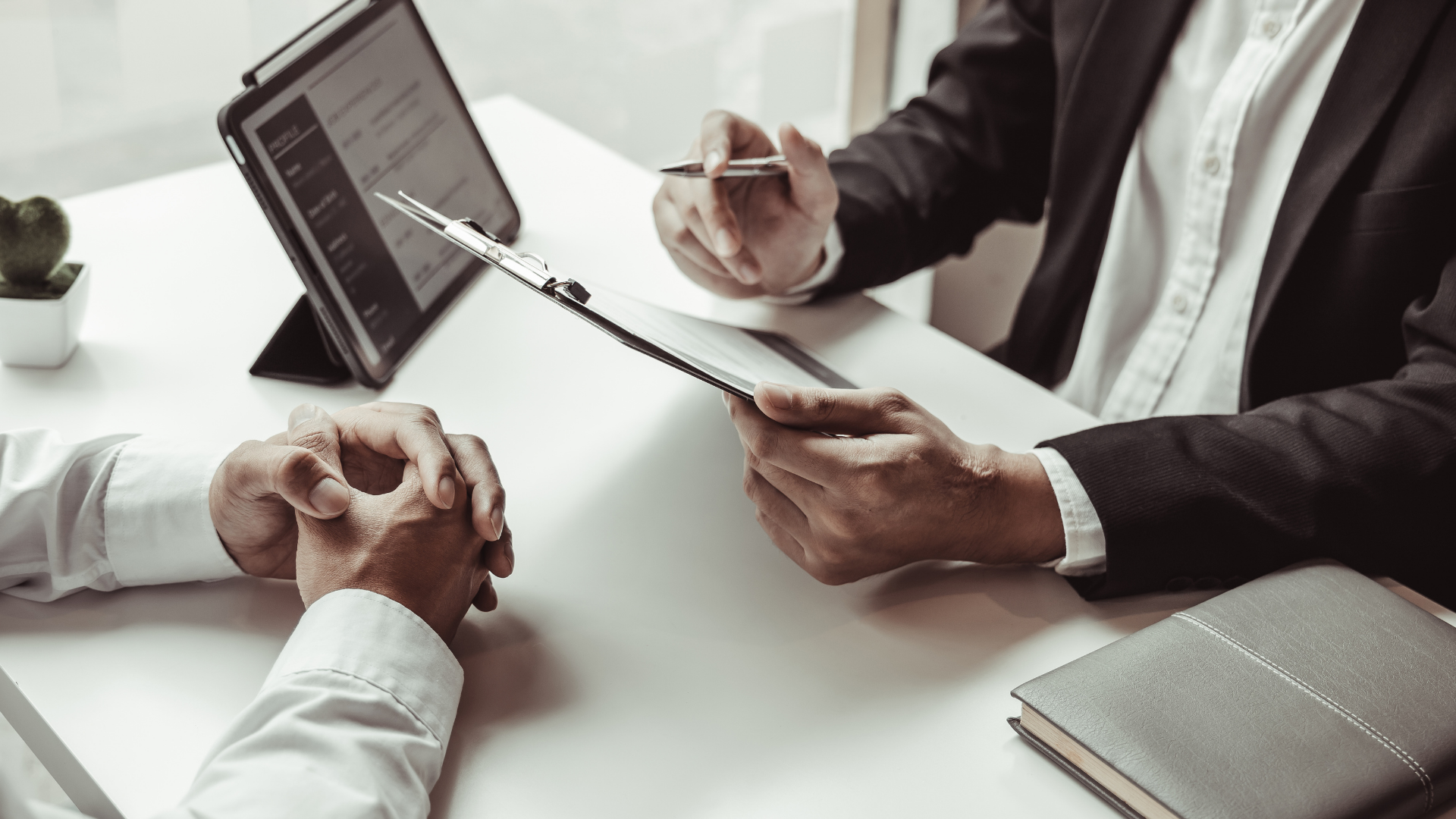Two professionals sit at a white desk during a formal meeting, reviewing documents and using a tablet.