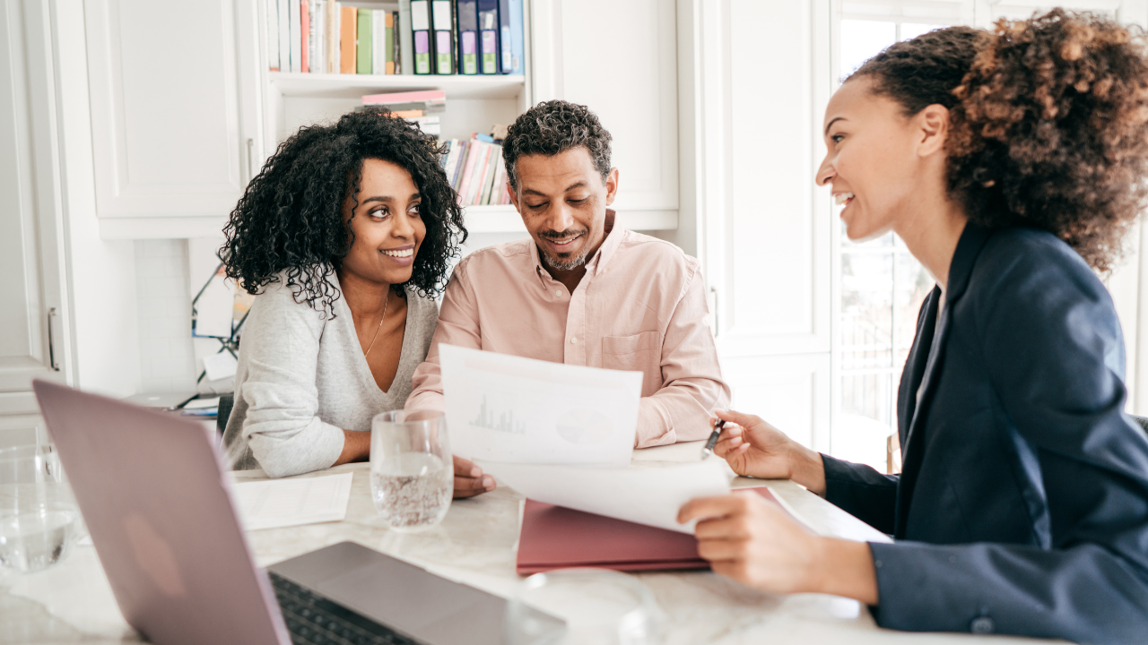 A consultant discusses paperwork with three people at a wooden table in a bright, modern office with plants.