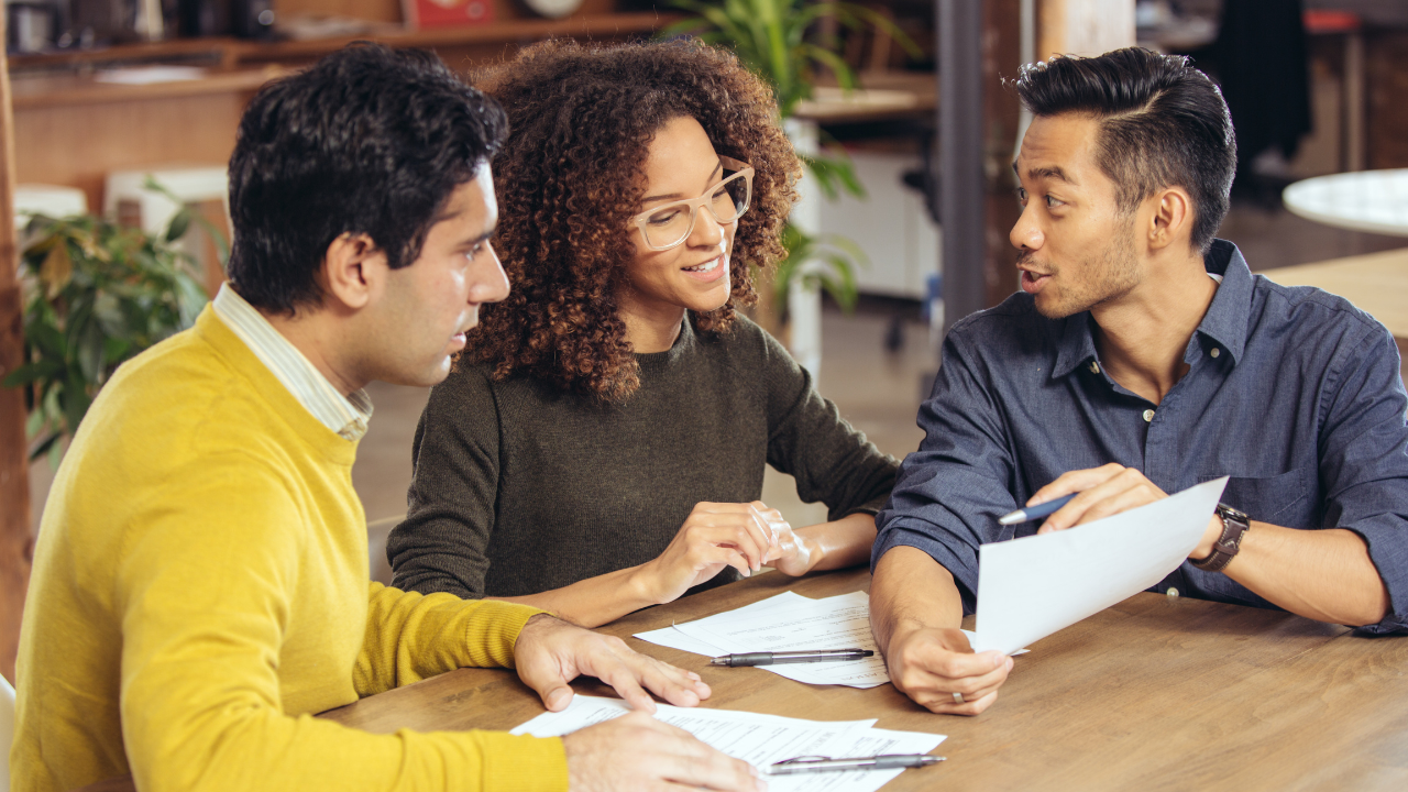 Three diverse people collaborating at a table, reviewing documents, and discussing ideas.