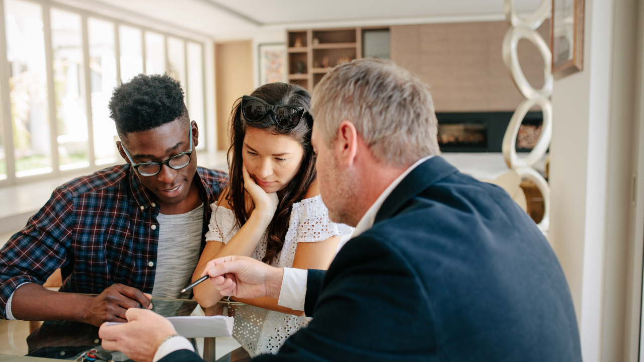 A professional in a suit explains a document to two people sitting at a table in a modern, well-lit office setting.