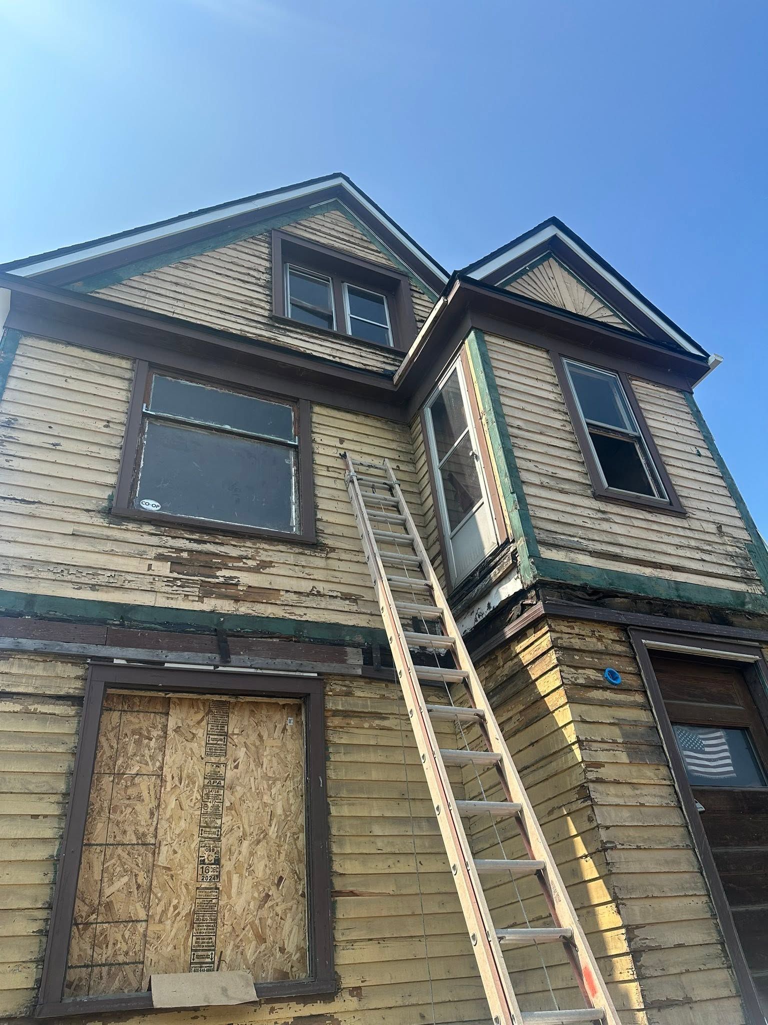 Boarded-up two-story house exterior with a ladder leaning to the upper floor under blue sky