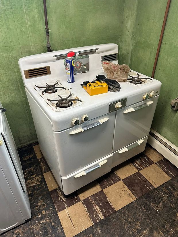White gas stove in a narrow utility space, with cleaning items and a bowl on top.