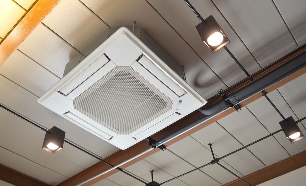 White ceiling-mounted HVAC unit with fan blades, surrounded by track lighting, against a cream-colored ceiling.