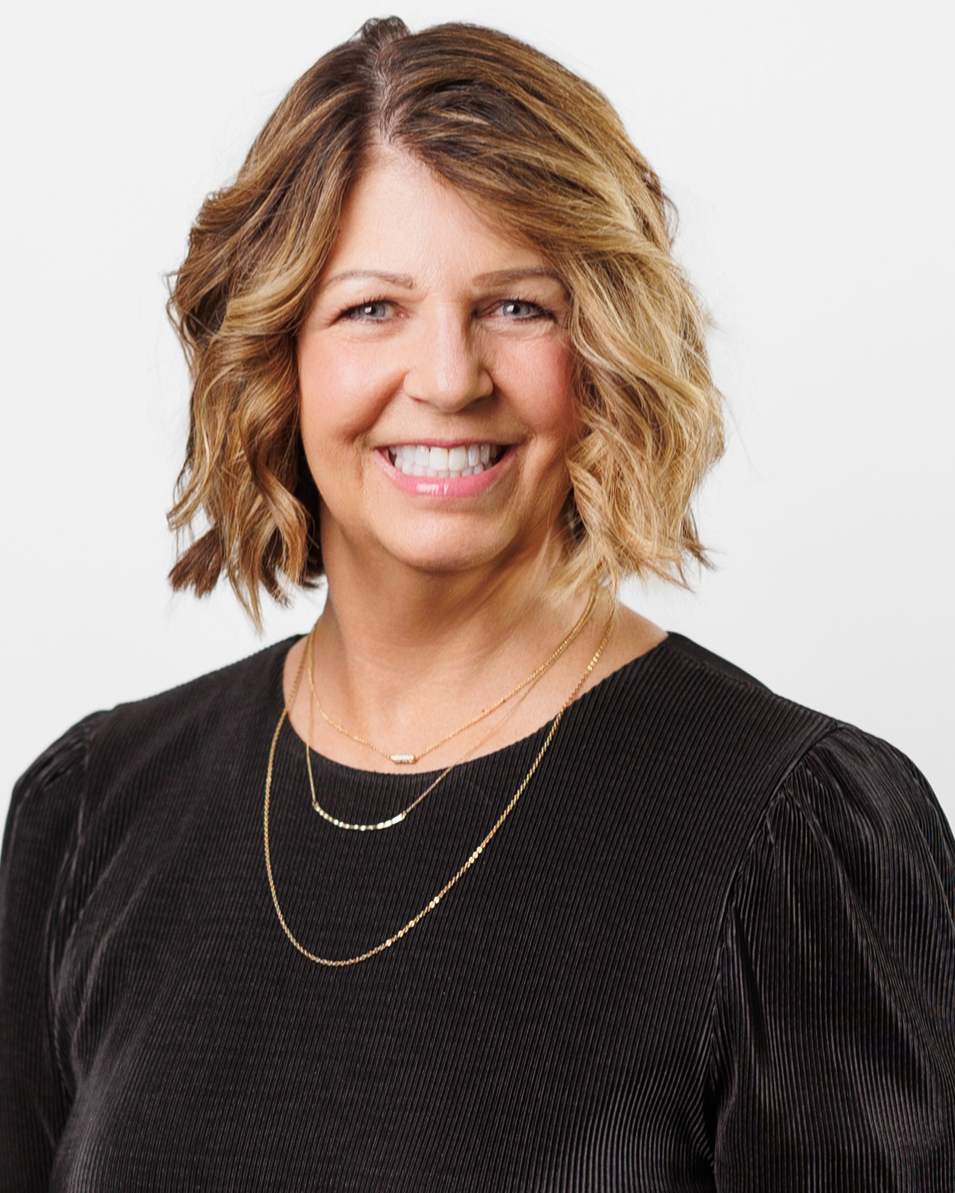 Woman with short, layered blonde hair smiles, wearing a black top and gold necklaces.