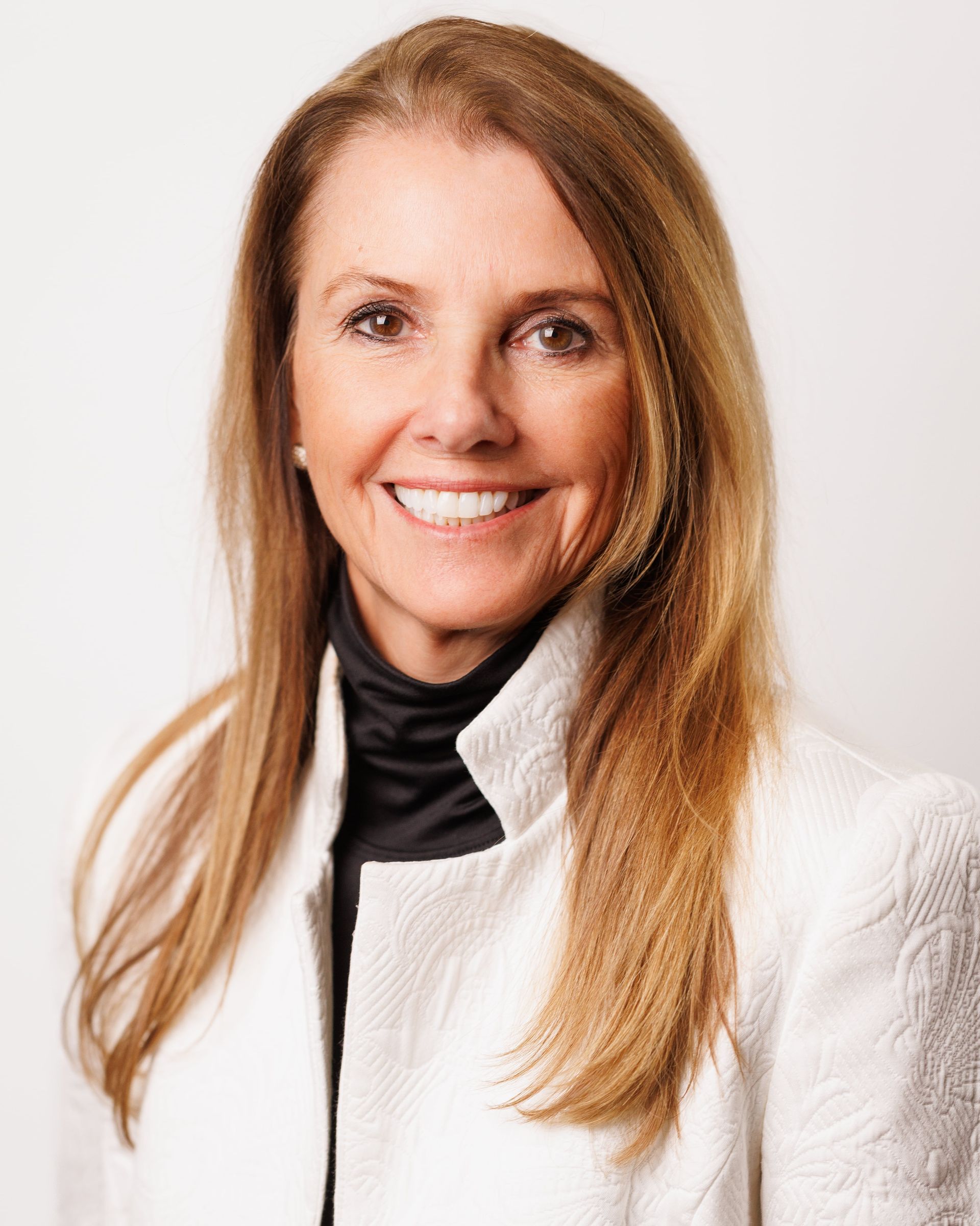 Woman with light skin wearing a white jacket over a black turtleneck, smiling at the camera.