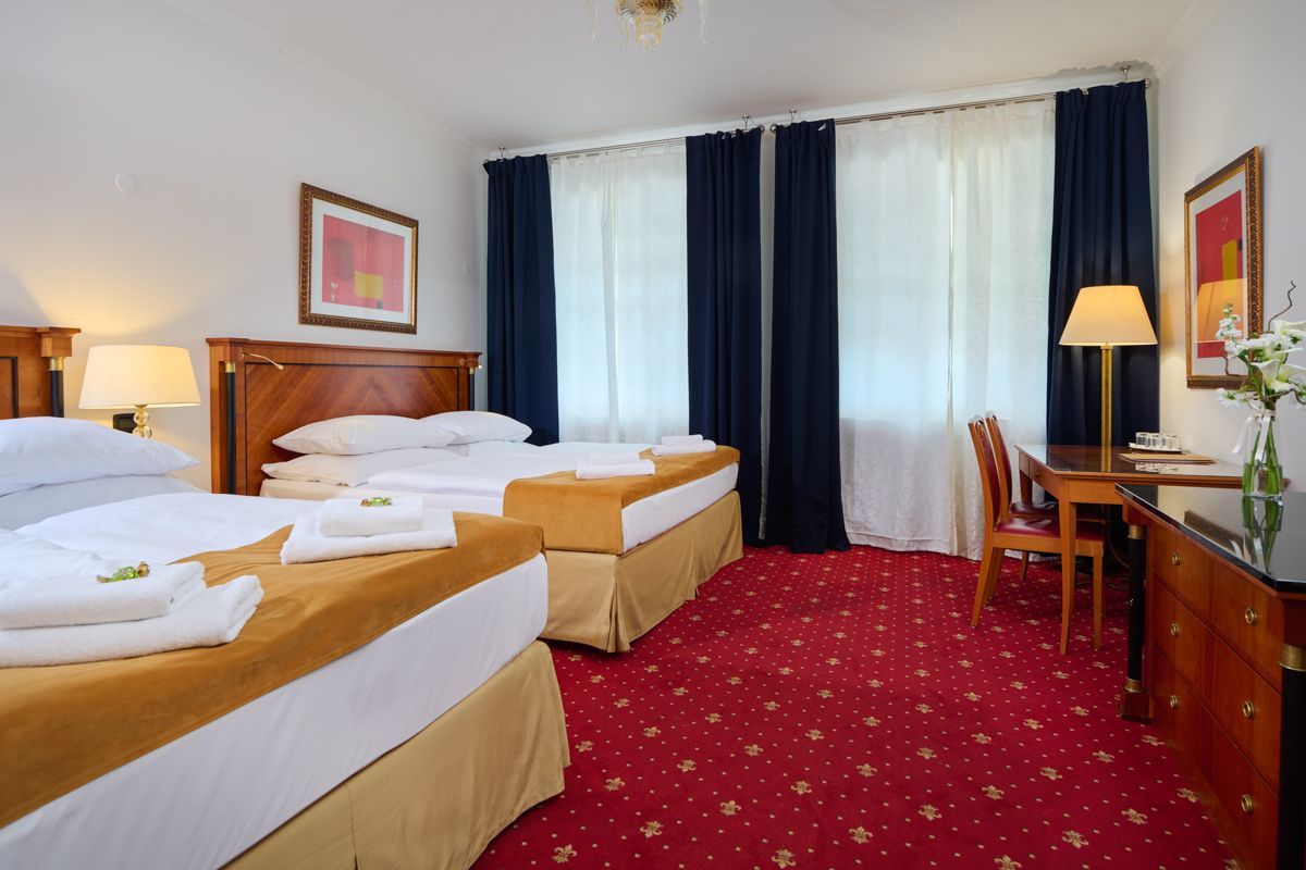 Camera d'albergo con due letti singoli, moquette rossa, scrivania e ampie finestre con tende blu.